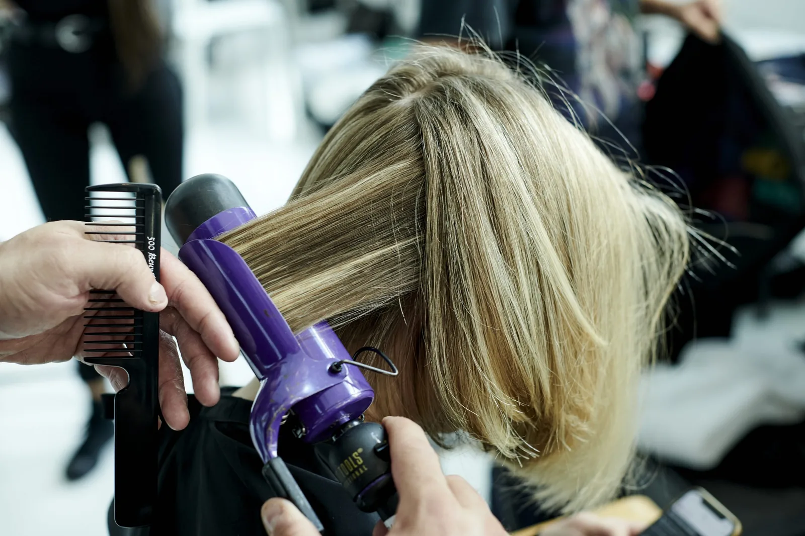 A stylist curling a client’s long hair with a wand at her station